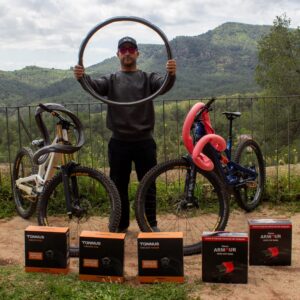 Bienve Aguado verkleed als Tannus houdt een ronde Tannus Tubeless insert vast, met zijn gezicht in het midden. Hij staat tussen twee van zijn fietsen en Tannus lekbeschermingsdozen.